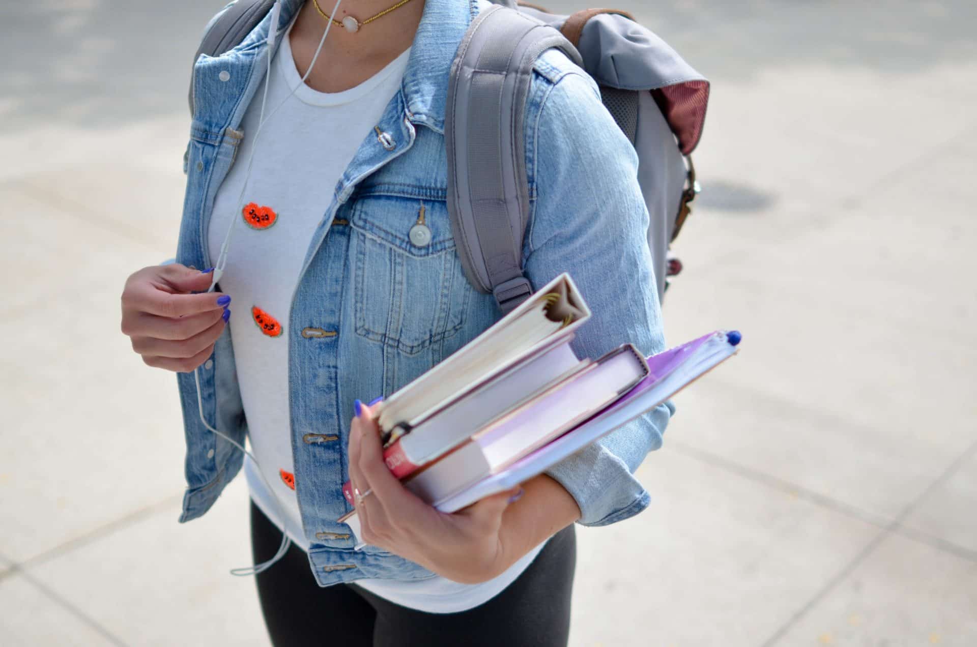 A picture of a person wearing a demin jacket with a rucksac on their back whilst holding a few books