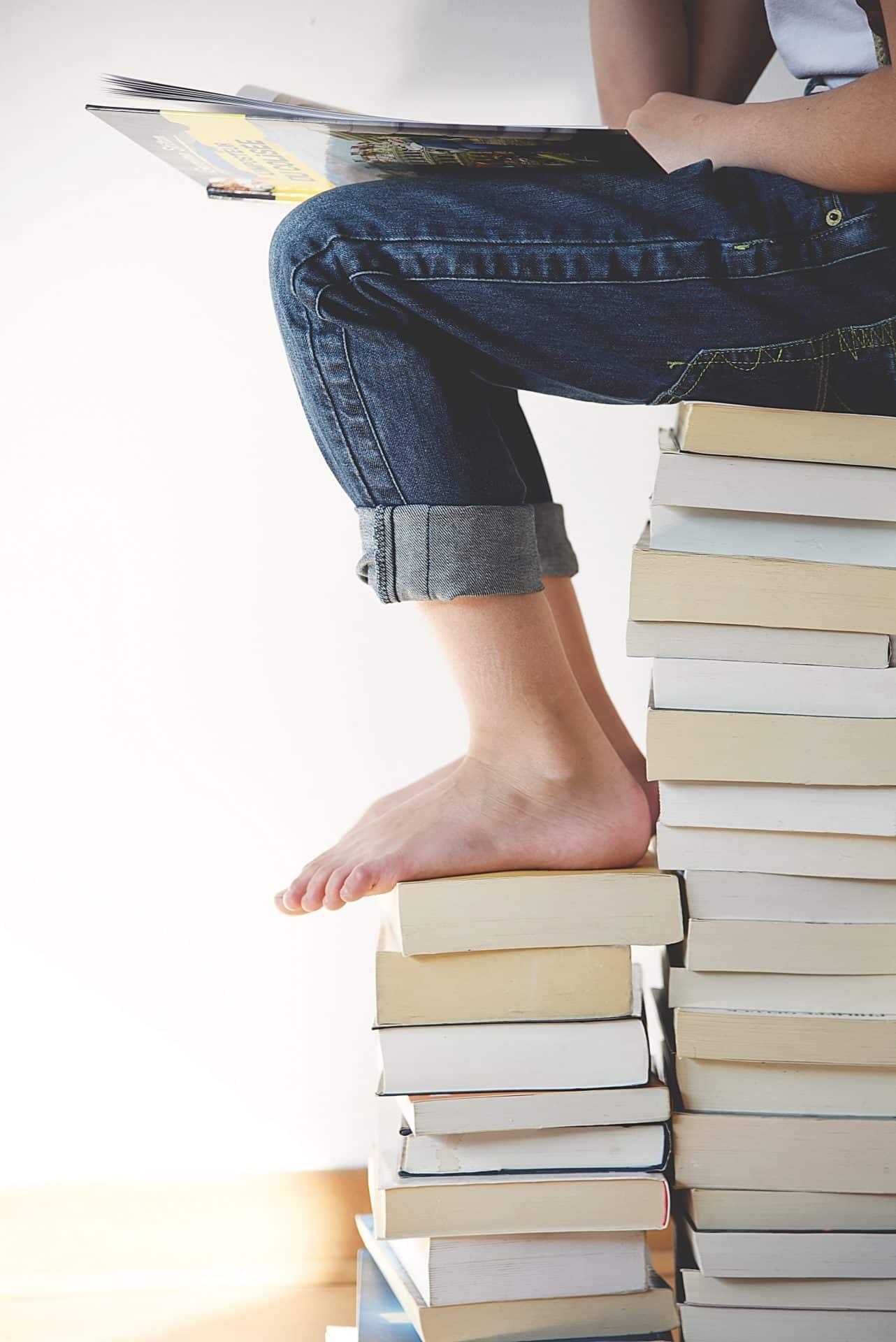 A persons legs. The person is sitting on lots of books stacked up