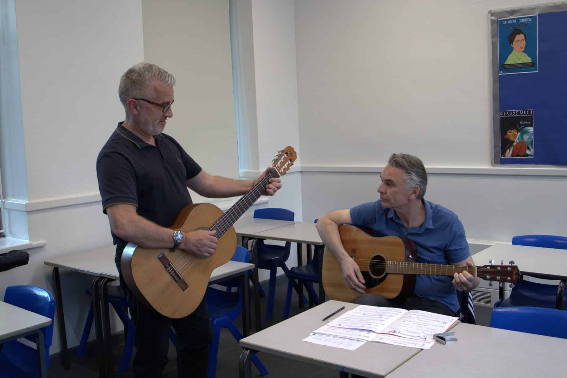 Two men (one sitting and one standing) playing the guitar