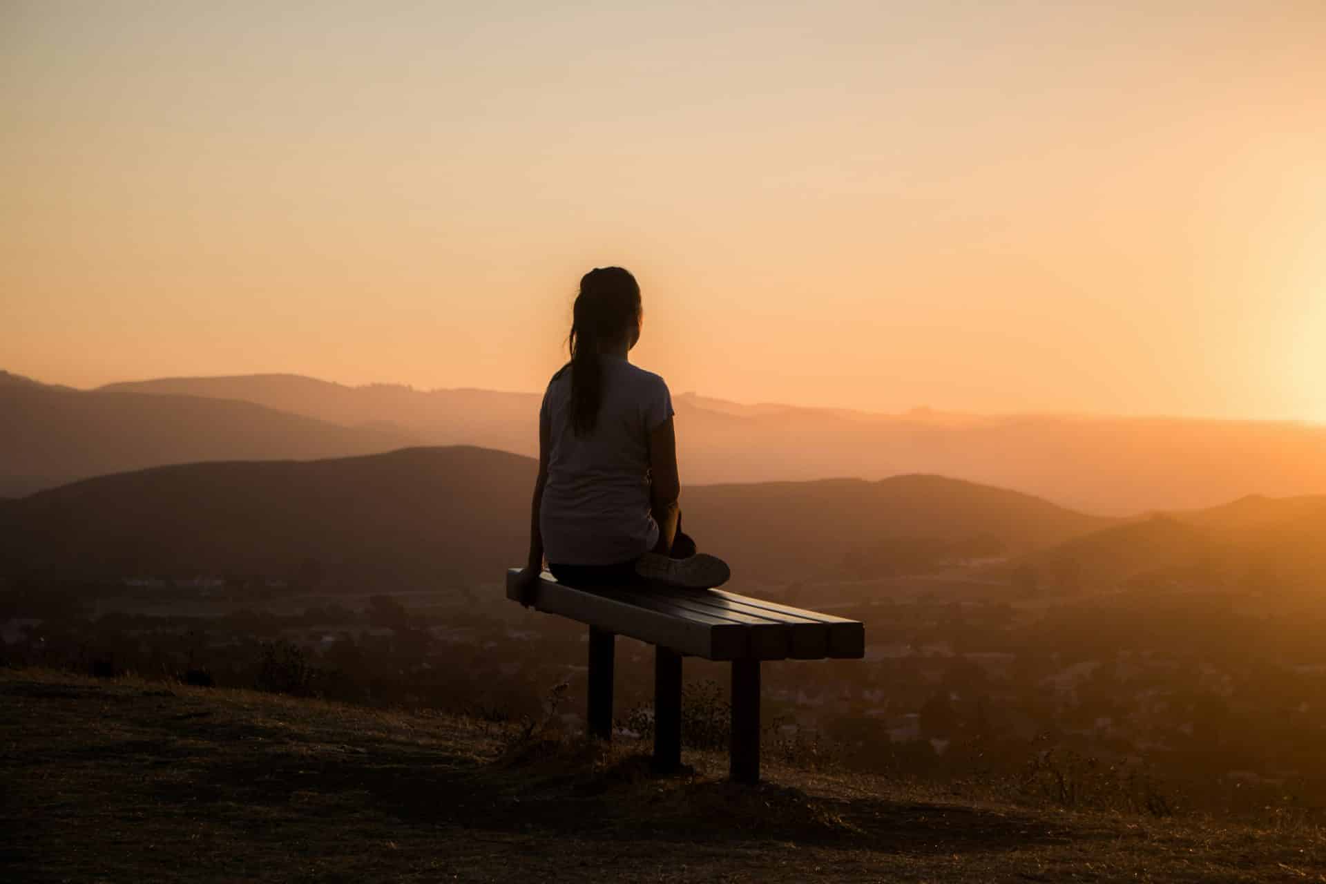A woman sitting with her legs up on a bench overlooking a beautiful sunset across hills