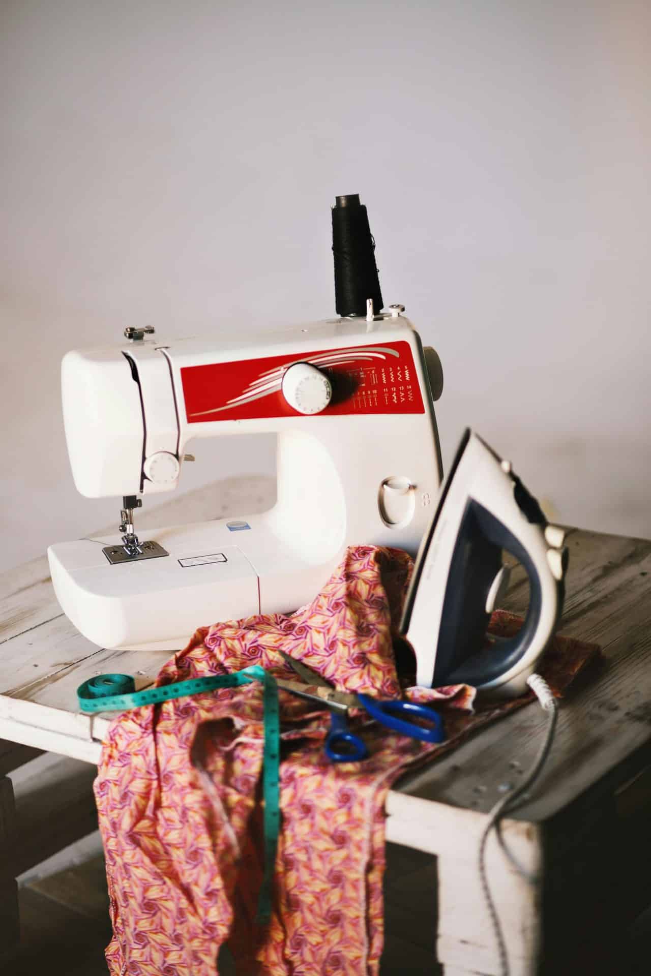A picture of a sewing machine, an iron, red fabric and a tape measure on a table