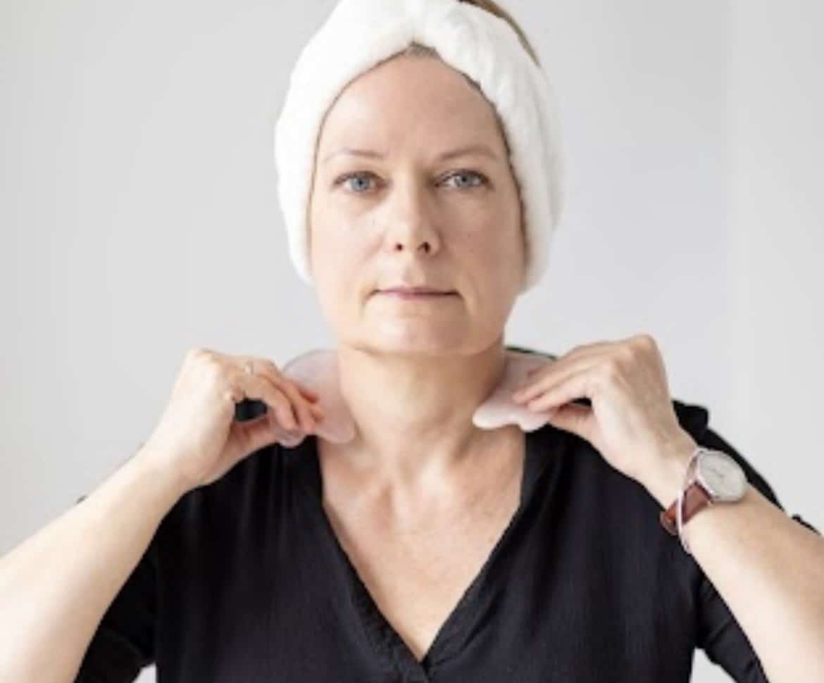 A woman wearing a hair turban, look at the camera whilst using gua sha tools on her neck