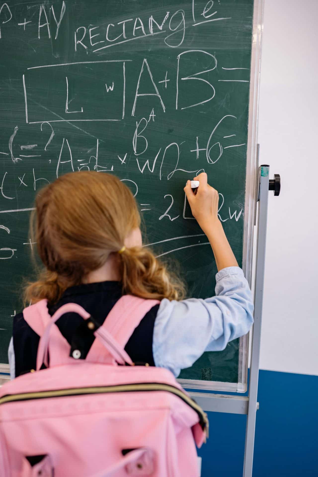 Child with Ginger hair and a pink rucksack on her back, standing at a chalk board, writing complicated sums