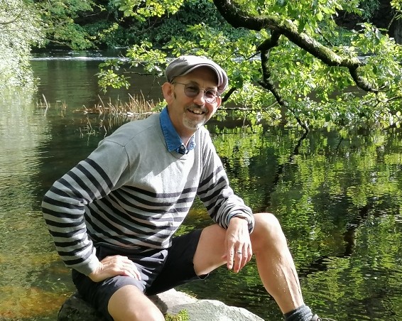 A relaxed man posing for a picture in a wooded area with a lake behind
